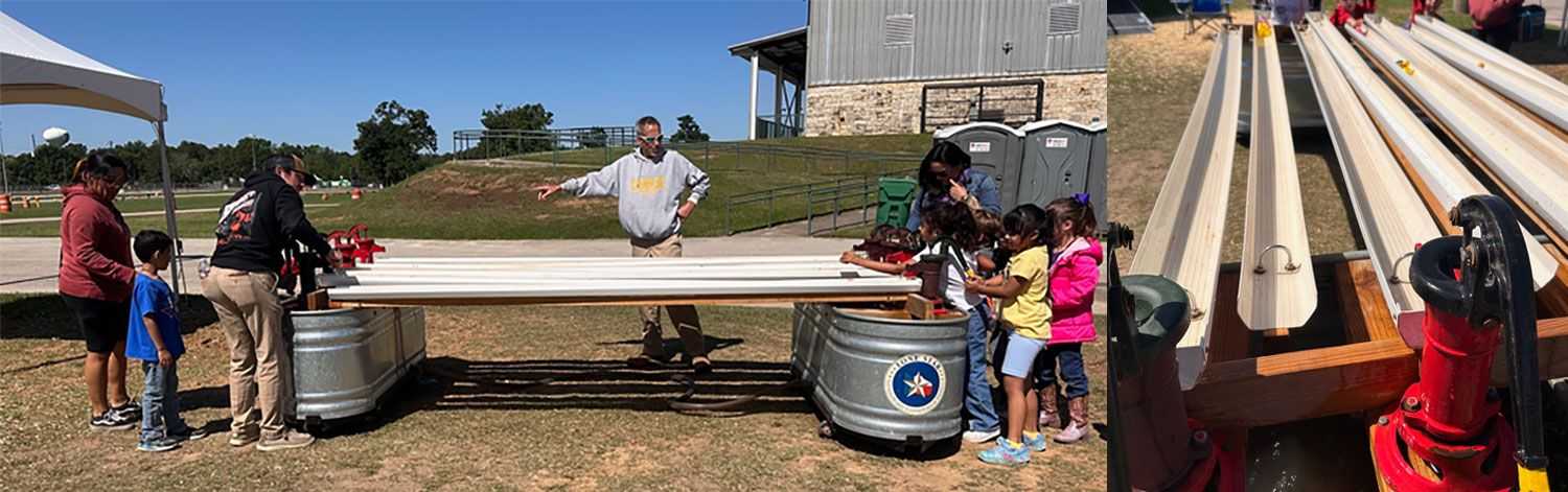 Children gather around a long, outdoor water trough activity featuring hand pumps and metal basins, while an instructor points toward the interactive display at a community event.